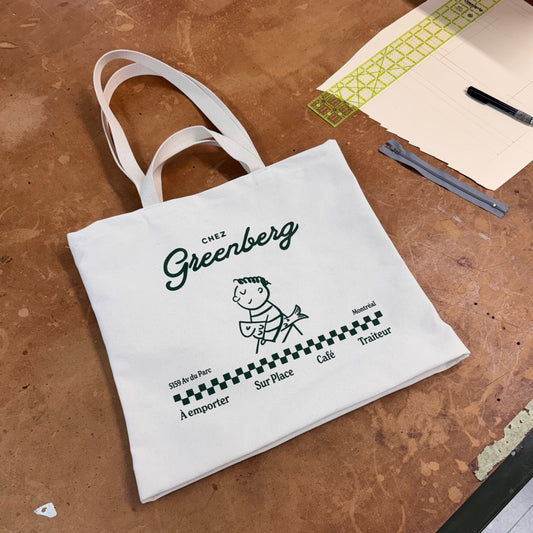 Chez Greenberg tote bag with both a shoulder strap and a hand strap. White canvas tote bag with green printed graphic of a kid and his fish.